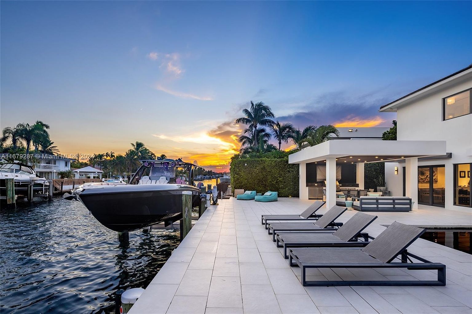 This image showcases a luxurious waterfront patio at sunset, featuring modern lounge chairs, a covered outdoor kitchen and dining area, and a sleek boat docked alongside. The architecture is contemporary with clean lines, and the overall impression is one of upscale relaxation and waterfront living. The camera is positioned at a medium angle, capturing the patio, boat, and the vibrant sunset sky.