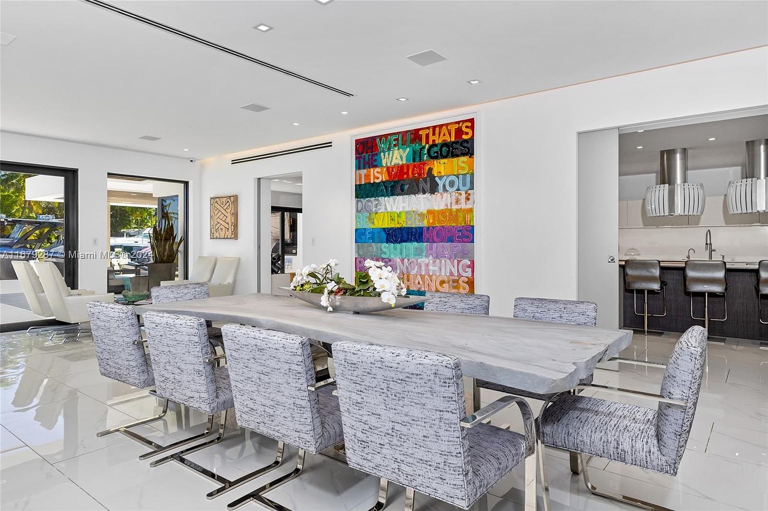 This interior shot showcases a modern dining room with a large, uniquely shaped dining table surrounded by upholstered chairs with metal frames. A colorful, framed artwork hangs on the wall, adding a vibrant focal point. The room features glossy tile flooring and is well-lit, creating a sophisticated and inviting atmosphere.