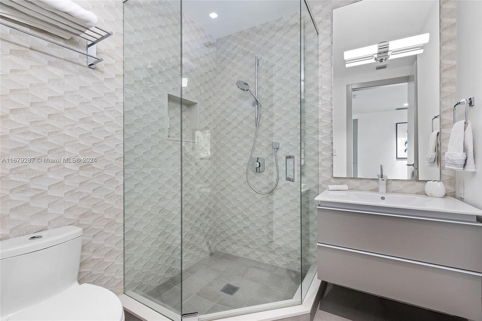 This is a well-lit bathroom featuring a modern design with textured geometric tiles on the walls. A glass-enclosed shower with a rainfall showerhead is visible, along with a floating vanity with a white countertop and gray drawers. A large mirror with integrated lighting enhances the space, creating a clean and contemporary feel.