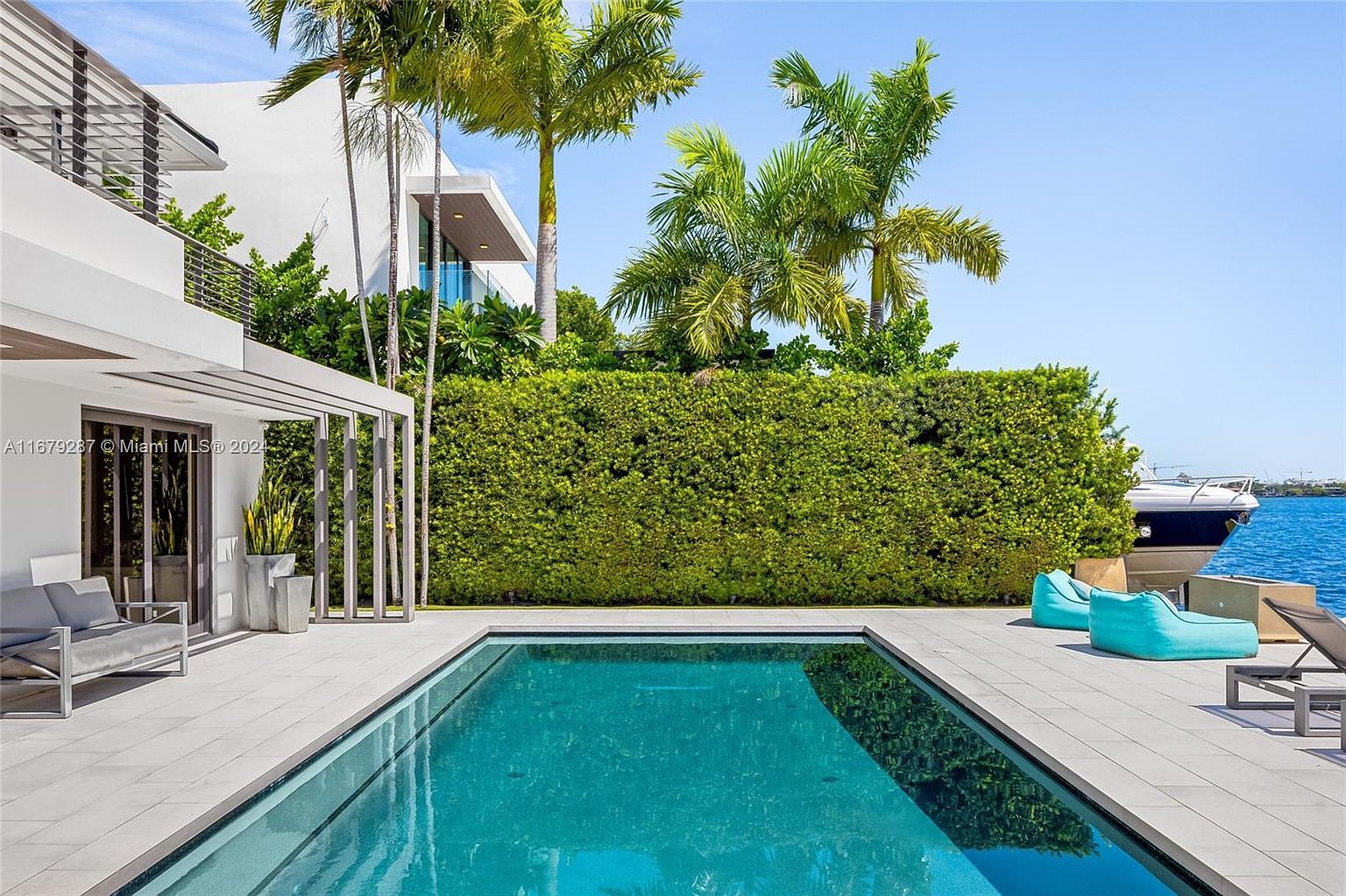 This image showcases a luxurious backyard pool area. The rectangular pool is surrounded by light gray tiling, with modern outdoor furniture including lounge chairs and beanbag chairs placed around the pool deck. A well-maintained green hedge provides privacy, and palm trees add a tropical feel, with a glimpse of a boat on the water in the background.