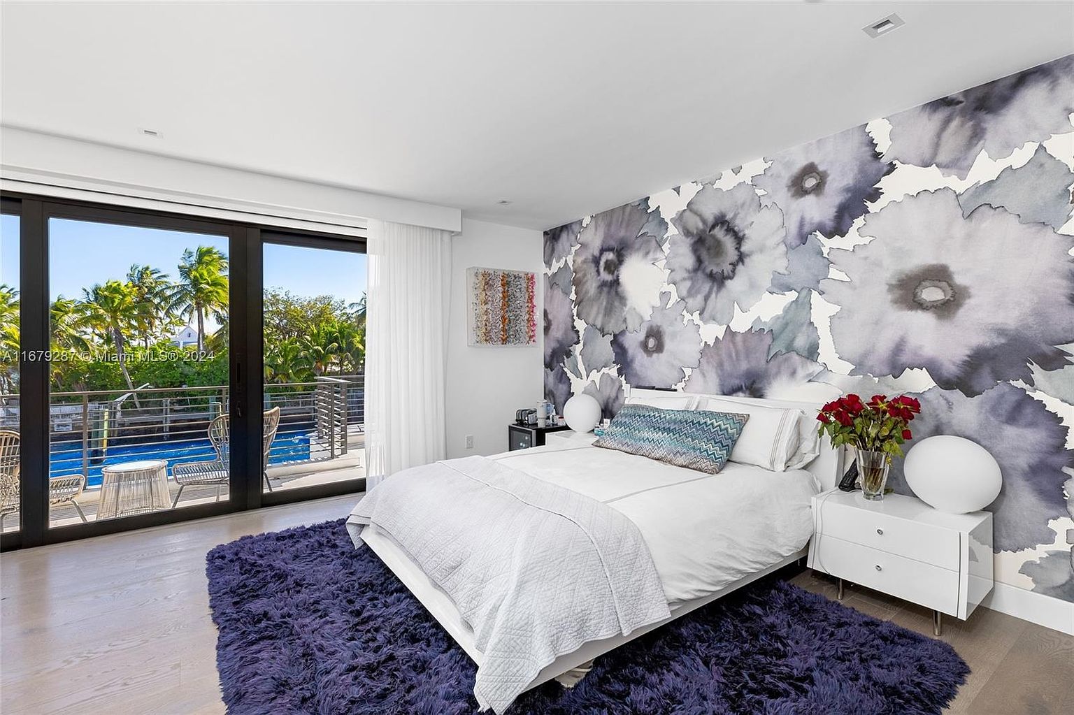 This is a primary bedroom featuring a king-size bed with white linens and a decorative blue pillow. The room has a modern aesthetic with a floral accent wall and a sliding glass door leading to a balcony overlooking a pool and lush greenery. A plush purple rug adds texture and warmth to the space.