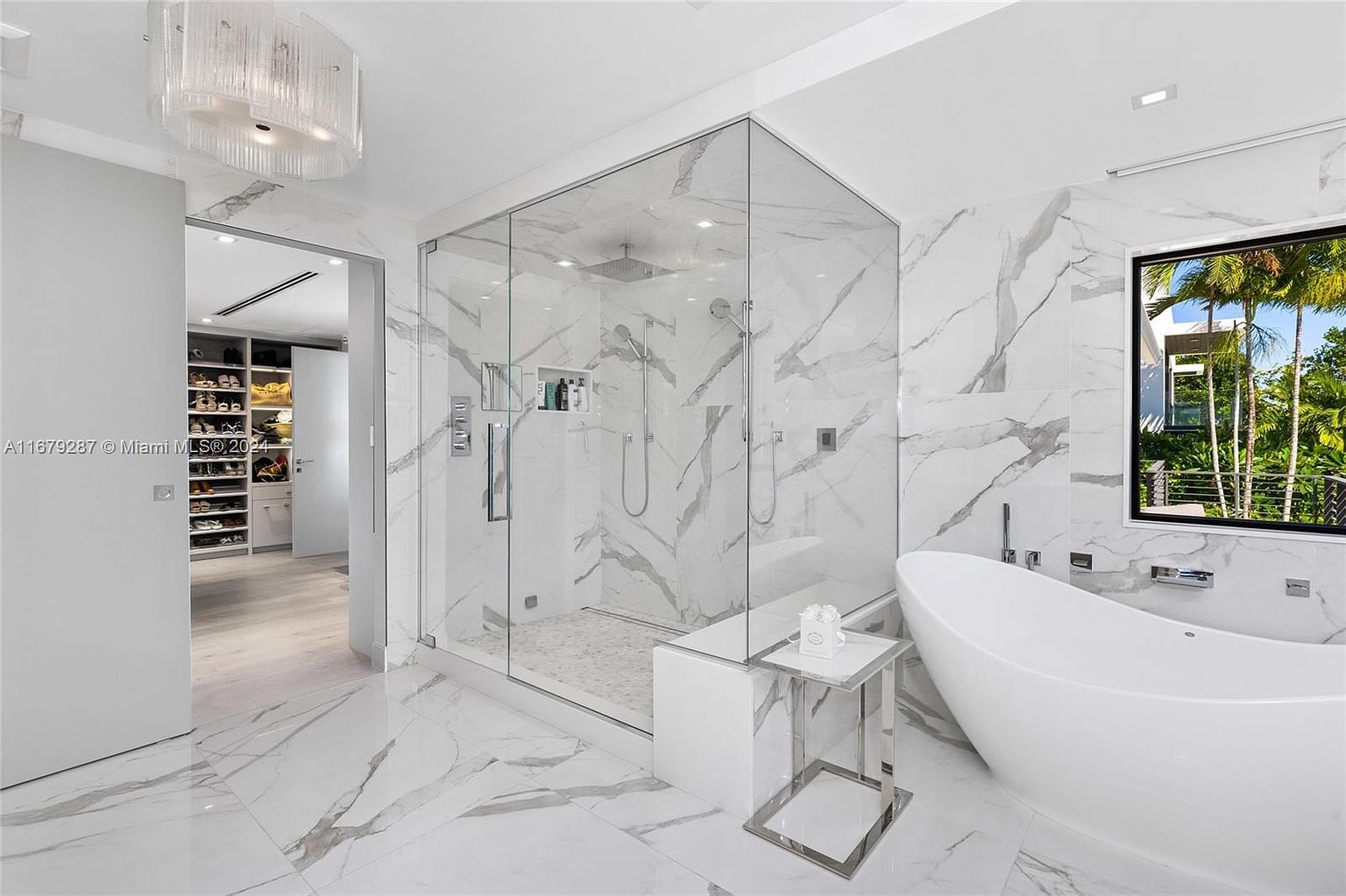 This is a luxurious primary bathroom featuring marble walls and flooring with striking gray veining. A glass-enclosed shower with dual shower heads and built-in shelving is a focal point, complemented by a freestanding white bathtub positioned near a window offering natural light and a view of lush greenery. A modern chandelier hangs above, adding to the room's elegance.