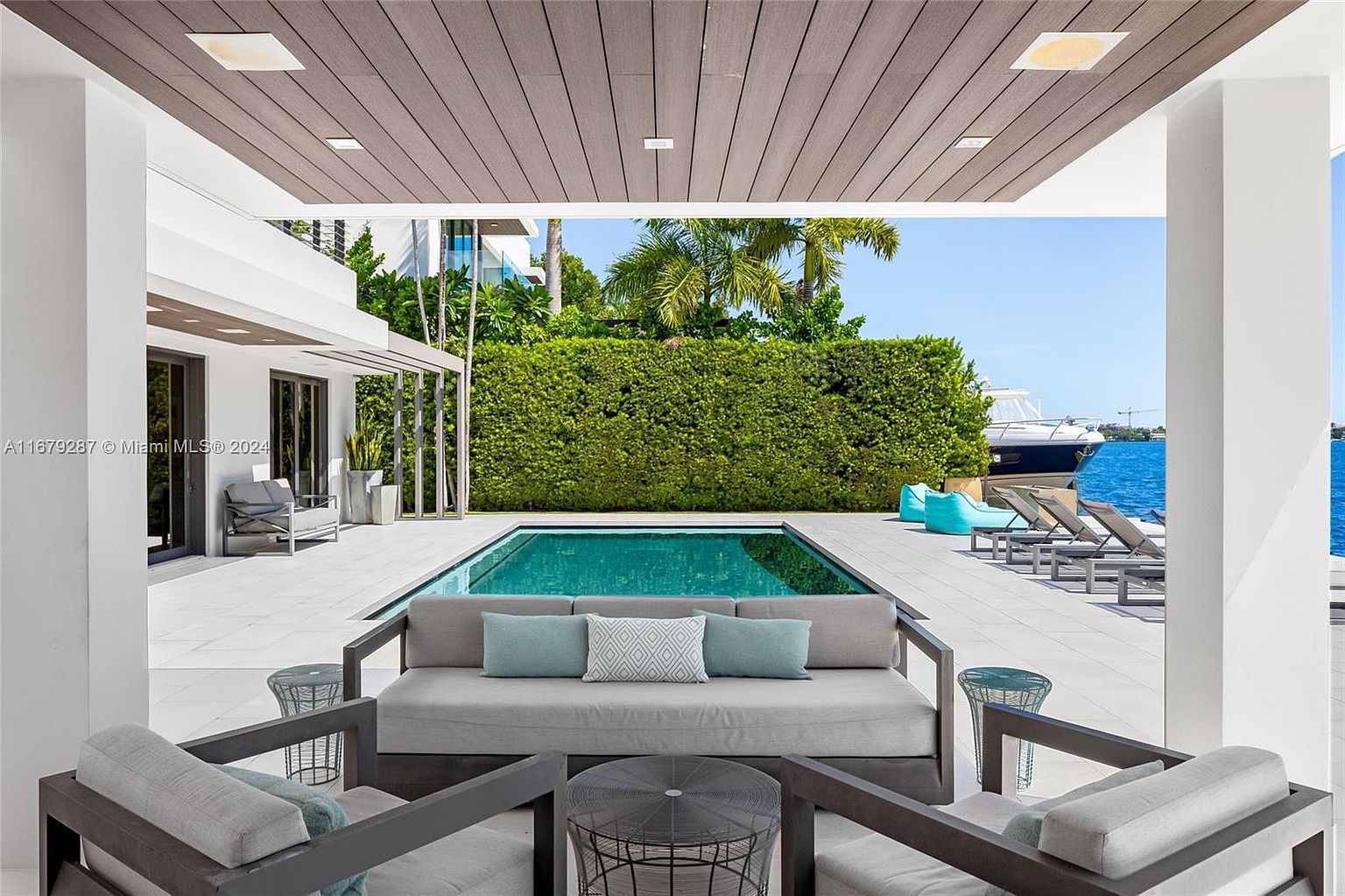 This image showcases a luxurious outdoor patio area with modern design elements. The space features comfortable lounge seating with stylish pillows, a sleek swimming pool, and a view of the waterfront with a boat docked nearby. The covered patio area provides shade and enhances the overall appeal of this outdoor living space.