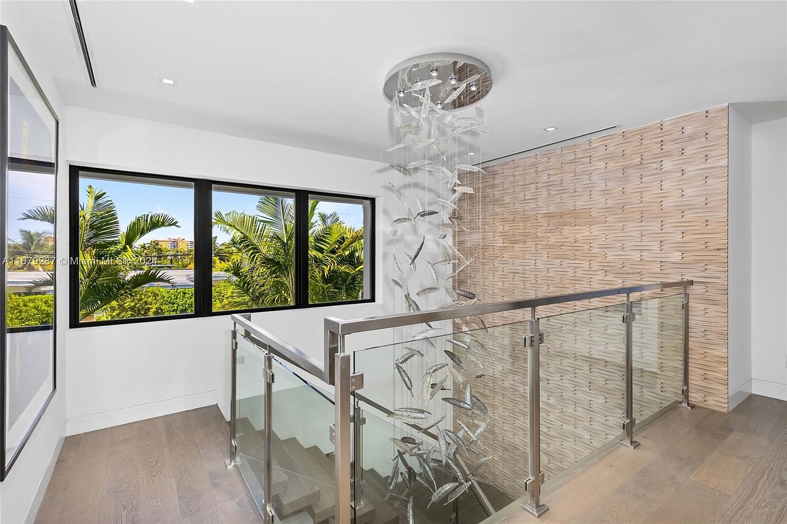 This interior shot showcases a modern hallway and staircase area, featuring a unique chandelier resembling falling leaves. The staircase is lined with glass and stainless steel railings, offering a sleek and contemporary design. A textured wall adds visual interest, while large windows provide natural light and views of lush greenery.