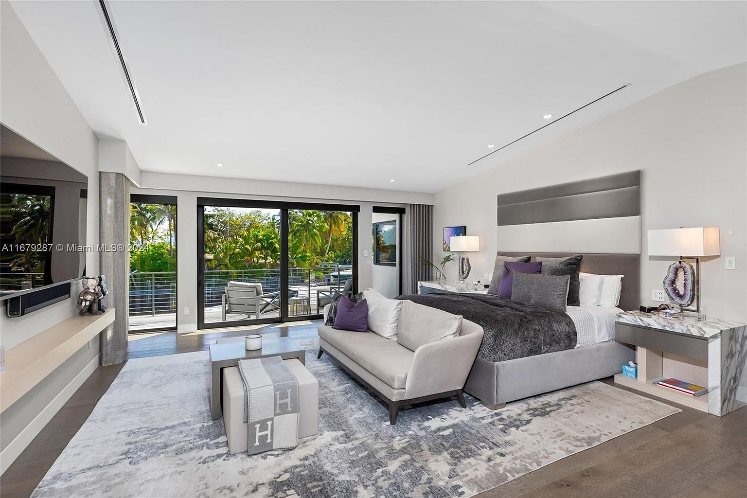 This is a luxurious primary bedroom featuring a king-size bed with a gray upholstered headboard and a plush gray comforter. The room includes a seating area with a stylish sofa and a coffee table, and large sliding glass doors open to a balcony with views of lush greenery. The color palette is primarily gray and white, creating a serene and sophisticated atmosphere.