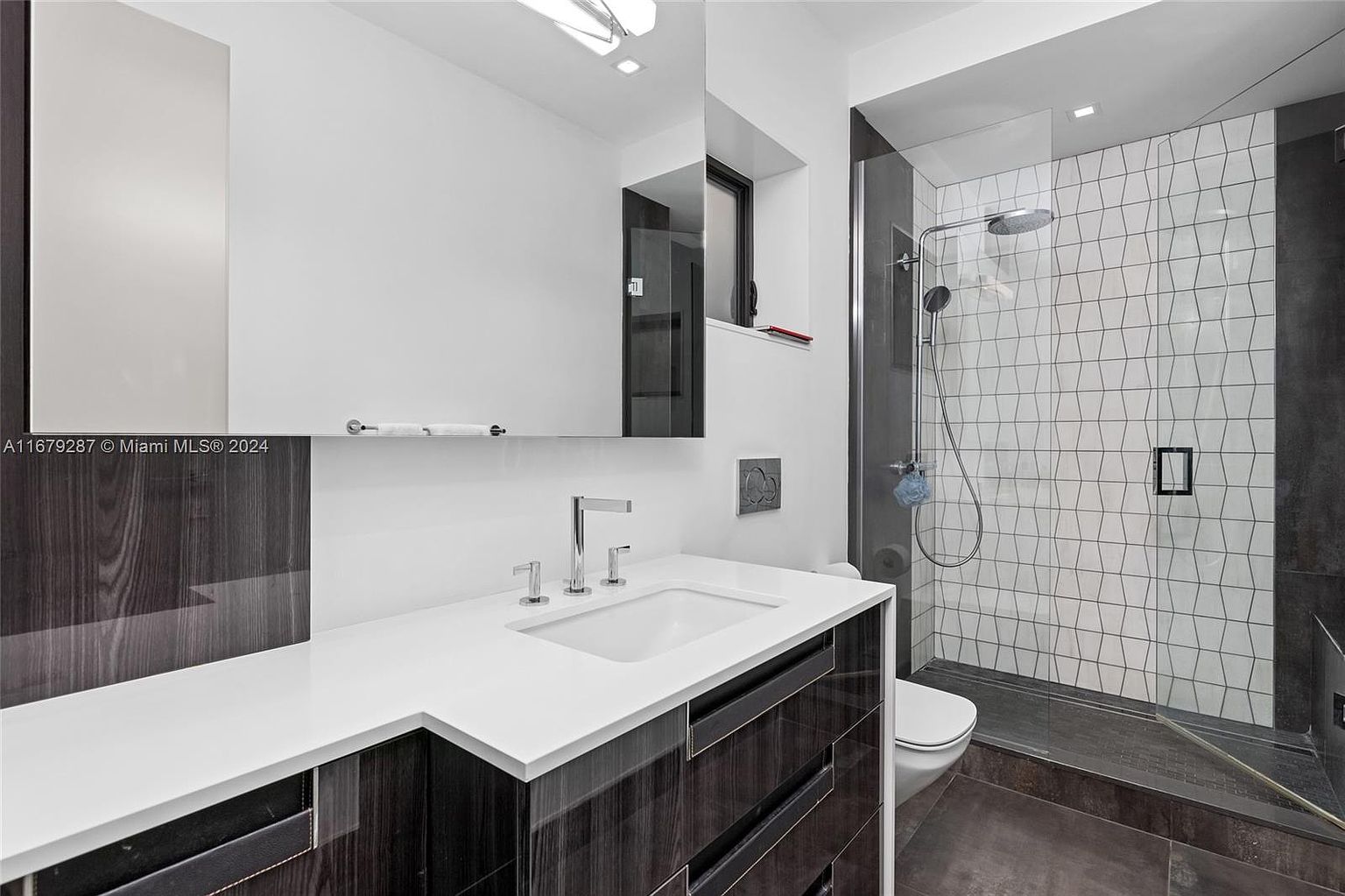 This is a modern bathroom featuring a sleek vanity with dark wood cabinetry and a white countertop. A large mirror hangs above the sink, and a glass-enclosed shower with geometric tilework is visible to the right. The overall design is minimalist and stylish, creating a clean and inviting space.