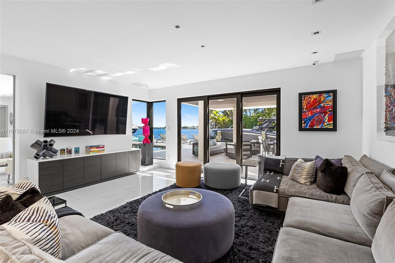 This is a modern living room featuring a large sectional sofa, a dark rug, and a large television mounted on the wall. Sliding glass doors offer a view of the outdoor pool and waterfront. The room is decorated with contemporary art and sculptures, creating a luxurious and inviting atmosphere.