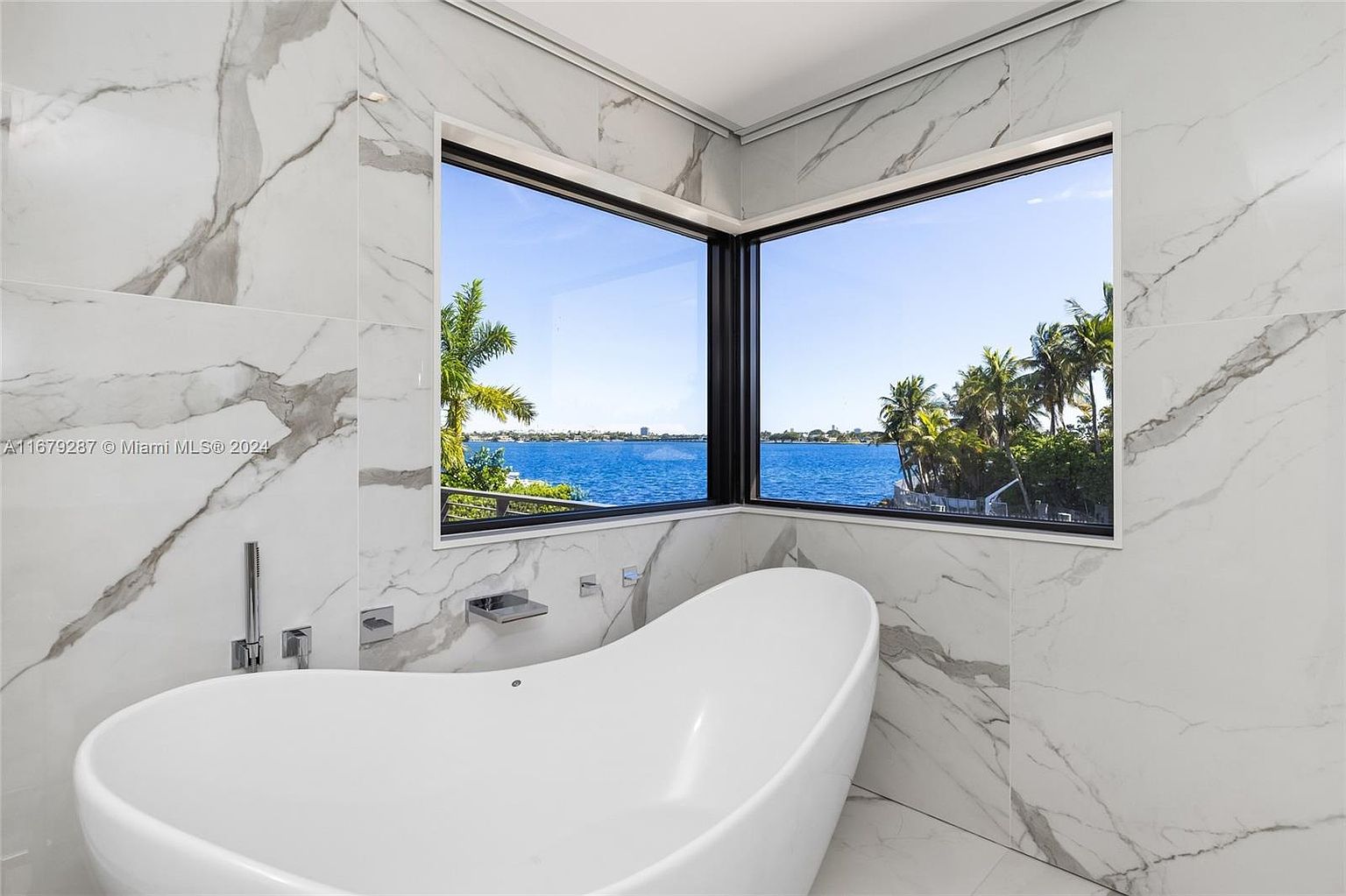 This is a luxurious primary bathroom featuring a freestanding white bathtub positioned in front of a large corner window with a view of the water and lush greenery. The walls and floor are covered in white marble with gray veining, creating a clean and elegant aesthetic. The chrome fixtures add a modern touch to the space.