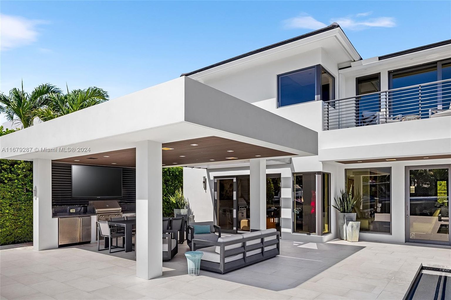 This image showcases a modern outdoor patio area with a covered outdoor kitchen and lounge space. The patio features sleek, gray outdoor furniture, a built-in grill, and a large flat-screen television, perfect for entertaining. The architecture is contemporary with clean lines, white columns, and a dark wood-paneled ceiling under the covered area, creating a luxurious and inviting outdoor living space.