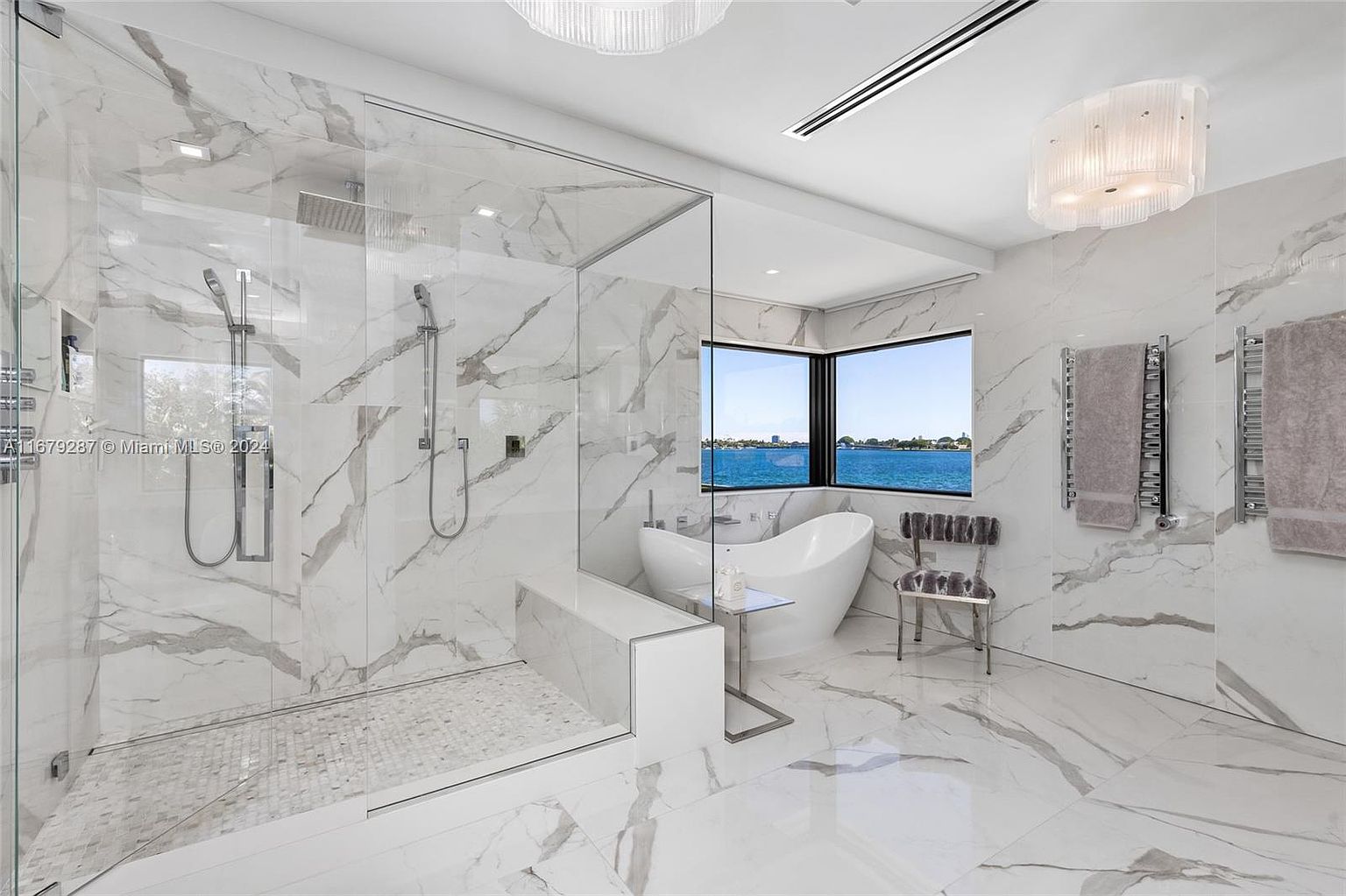This is a luxurious primary bathroom featuring marble walls and flooring, a glass-enclosed shower with dual showerheads, and a freestanding soaking tub positioned in front of a large window with a water view. The bathroom also includes heated towel racks and a stylish chair, creating a spa-like atmosphere. The perspective is wide, showcasing the spaciousness and high-end finishes.