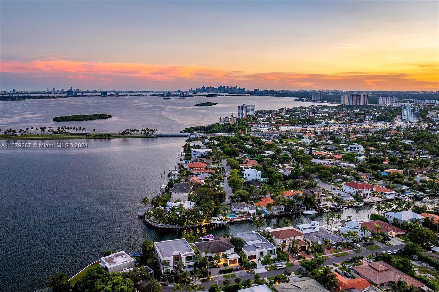This aerial view showcases a luxurious waterfront neighborhood at sunset. The scene features upscale homes with pools and boat docks along canals, lush greenery, and a distant cityscape skyline. The warm colors of the sky reflect on the water, creating a serene and inviting atmosphere.