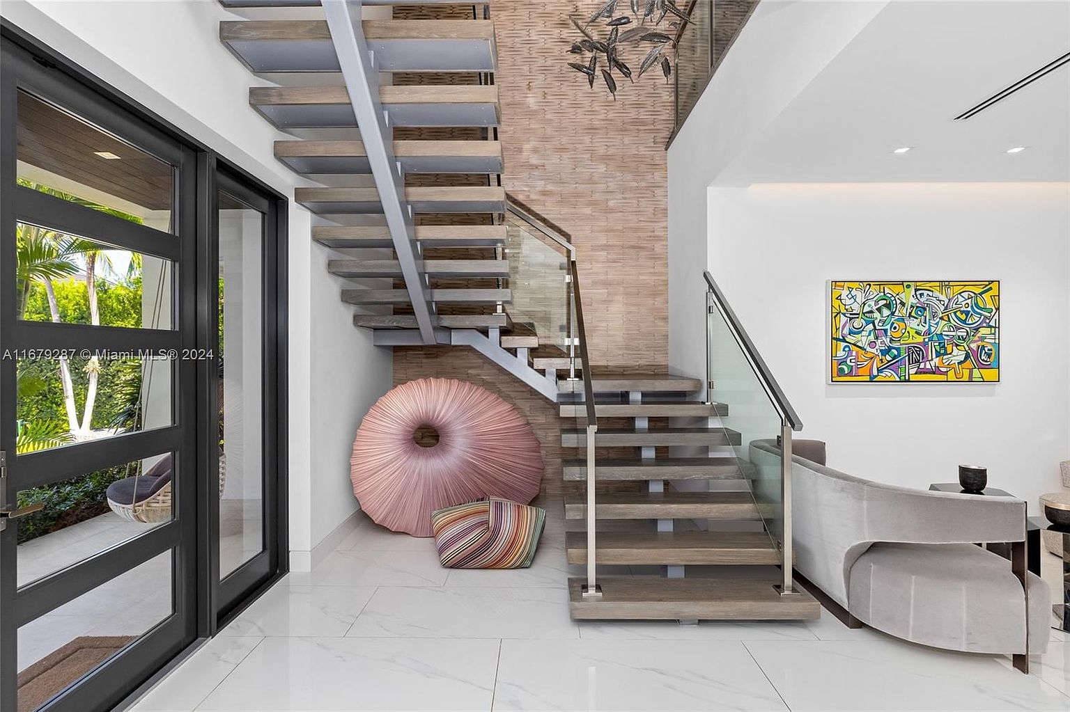 This interior shot showcases a modern hallway with a striking staircase design. The floating stairs feature wooden treads and glass railings, complemented by a textured wall. A large, round pink cushion and a striped pillow add a touch of whimsy, while an abstract painting enhances the contemporary aesthetic.