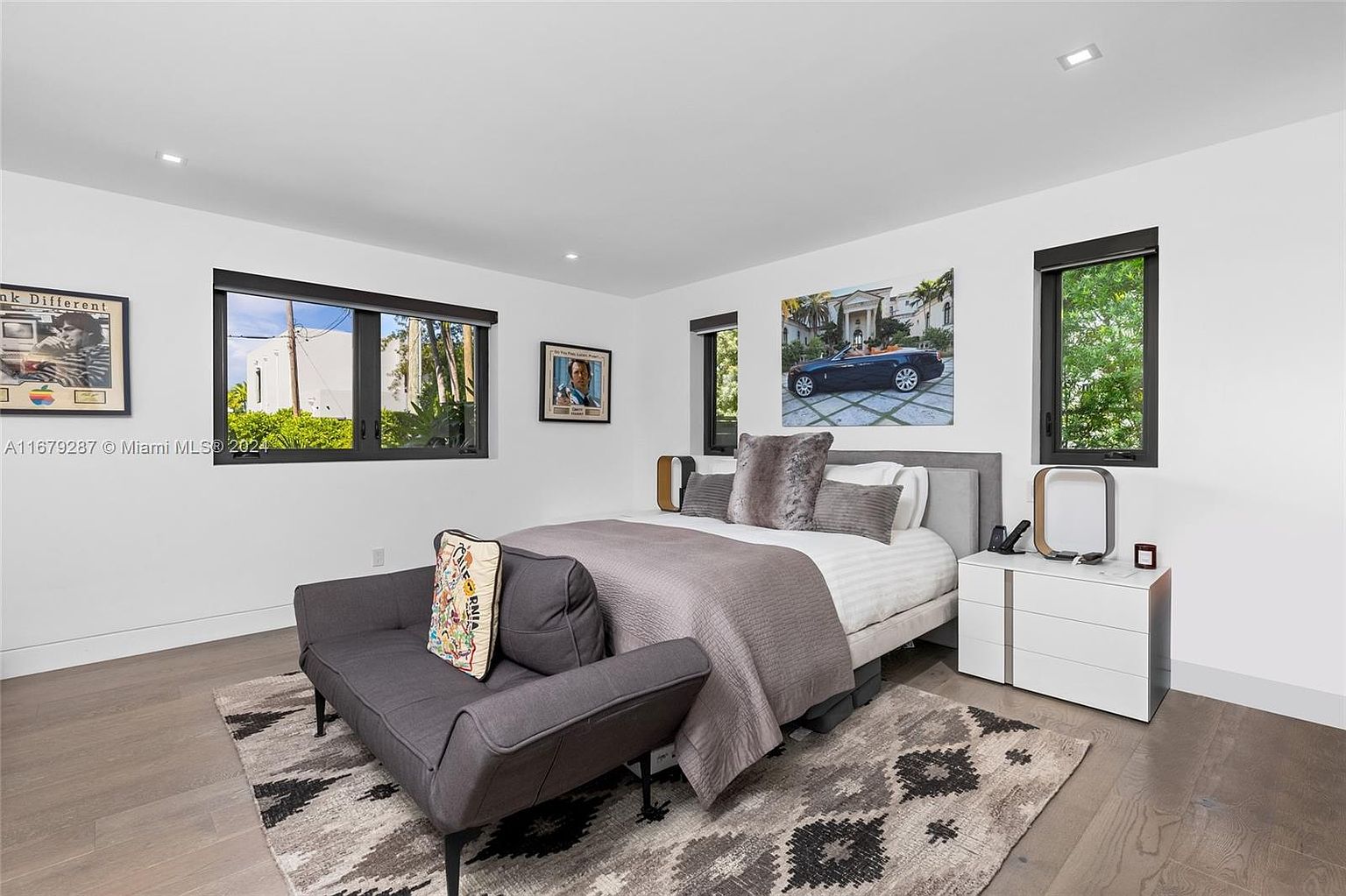 This is a well-lit primary bedroom featuring a neutral color palette with white walls and light wood flooring. The room includes a bed with a gray headboard and bedding, a gray settee, a white nightstand, and several framed pictures on the walls. A patterned rug adds texture to the space, and natural light streams in through the windows.