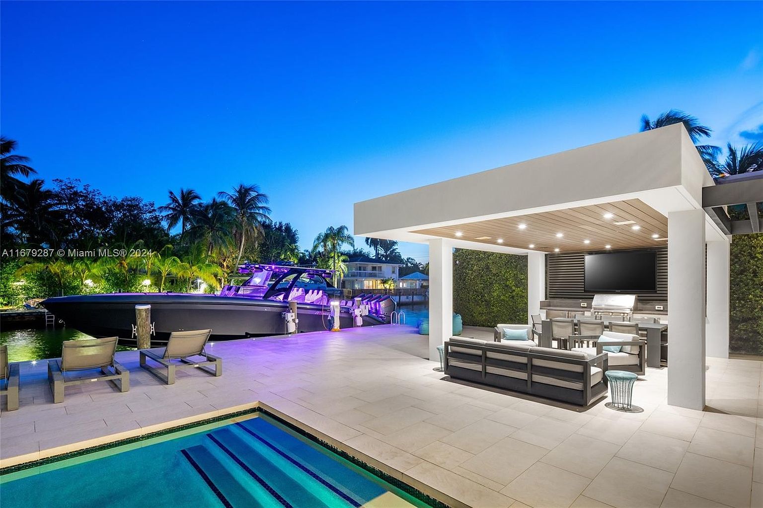 This image showcases a luxurious outdoor living space with a modern patio, a covered outdoor kitchen and dining area, and a sleek swimming pool. A large boat is docked nearby, adding to the upscale ambiance. The scene is set against a twilight sky, highlighting the property's waterfront location and sophisticated design.
