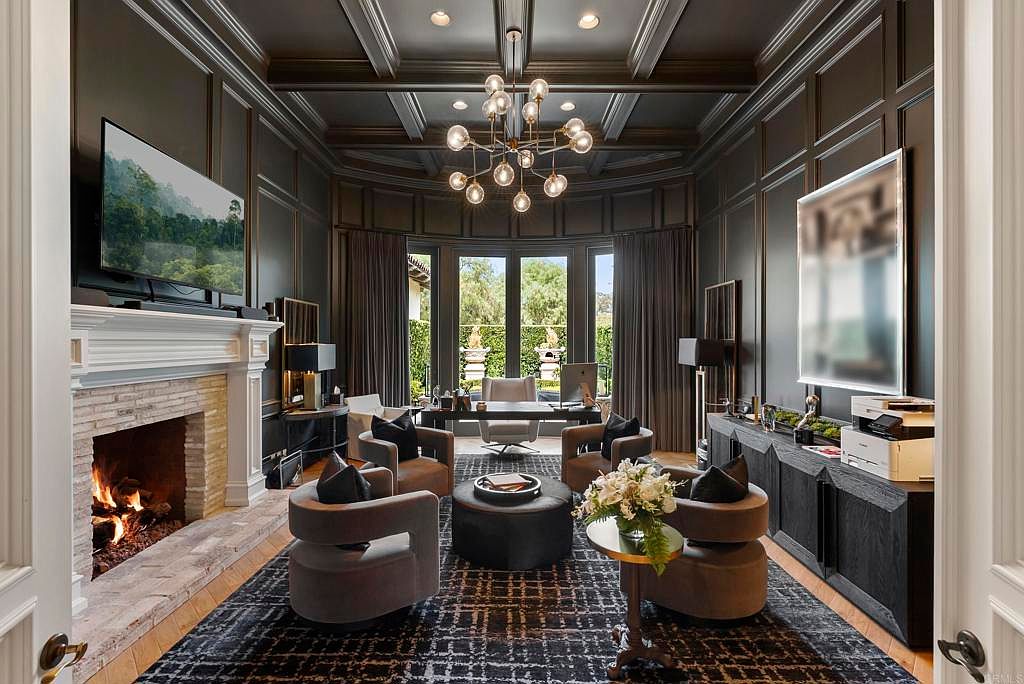 This is a luxurious home office featuring dark wood paneling, a coffered ceiling with a modern chandelier, and a fireplace. The room is furnished with comfortable armchairs, a central ottoman, and a desk positioned in front of a large window offering natural light and a view of the exterior. The overall impression is one of sophistication and comfort, ideal for a high-end real estate listing.
