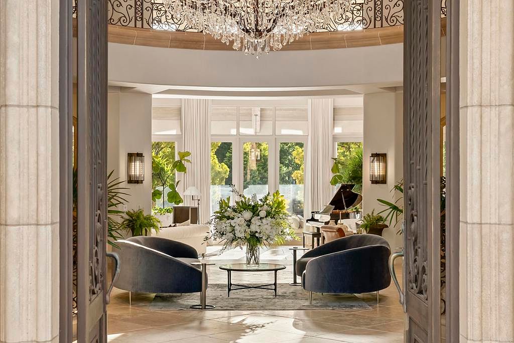 This is an interior shot of a luxurious living room, viewed through an ornate doorway. The room features elegant furnishings, including two plush armchairs, a glass-topped coffee table with a floral arrangement, and a grand piano in the background. Large windows provide ample natural light, and a crystal chandelier adds a touch of opulence.