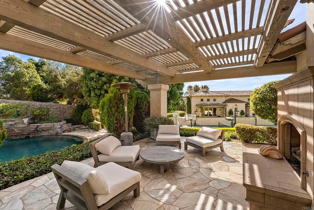This image showcases a luxurious outdoor patio area, featuring comfortable lounge seating arranged around a round coffee table. The patio is covered by a wooden pergola, providing shade and an elegant architectural detail. A stone fireplace adds warmth and character, while the surrounding landscaping, including a pool and lush greenery, creates a serene and inviting atmosphere.