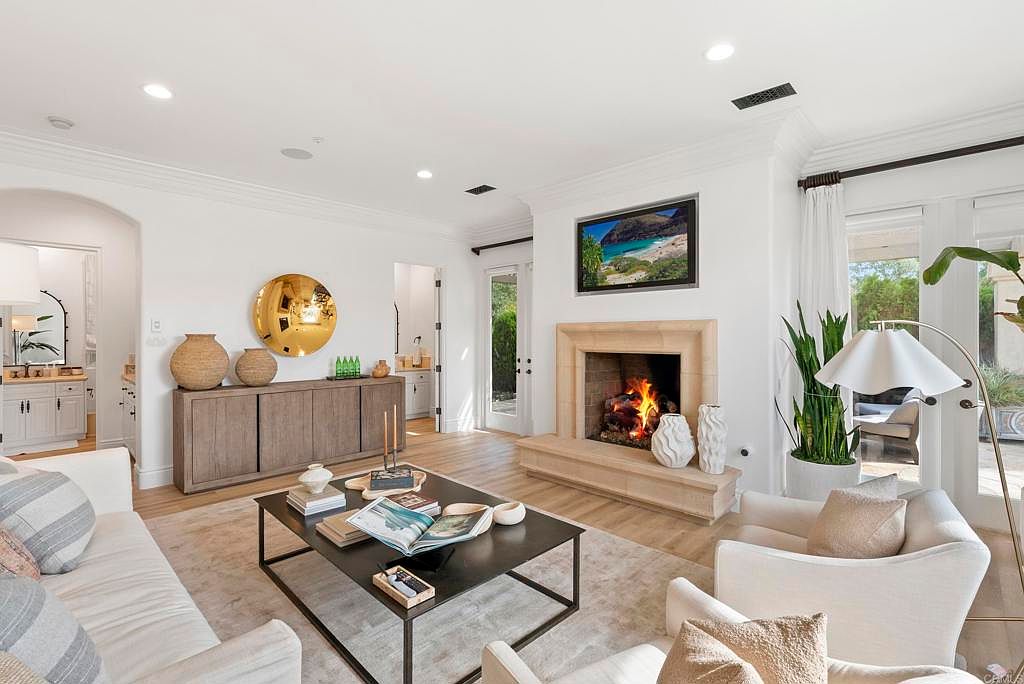 This is an interior shot of a living room featuring a fireplace with a TV mounted above it. The room is decorated in neutral tones with white sofas, a dark coffee table, and a light-colored rug. A large gold mirror and wooden cabinet are on the left wall, and natural light streams in from the windows and doors on the right, creating a bright and inviting atmosphere.