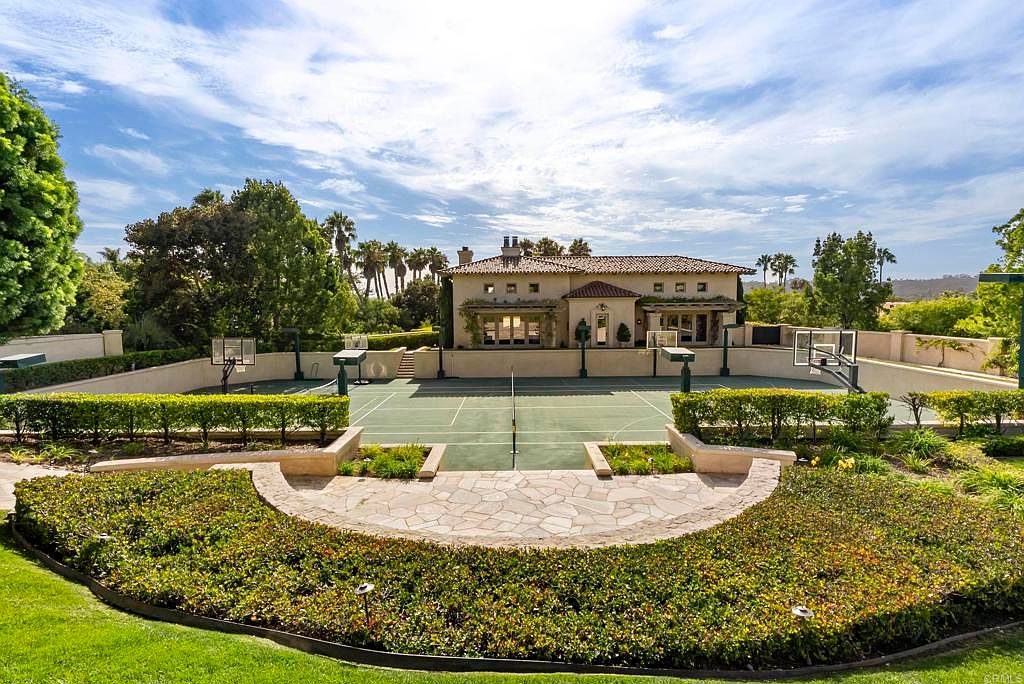 This image showcases a beautifully landscaped yard and garden area, featuring manicured hedges, stone pathways, and a well-maintained lawn. In the background, a large house with a tile roof overlooks a tennis court, creating a luxurious and private outdoor space. The scene is bathed in sunlight, highlighting the lush greenery and the overall tranquility of the property.