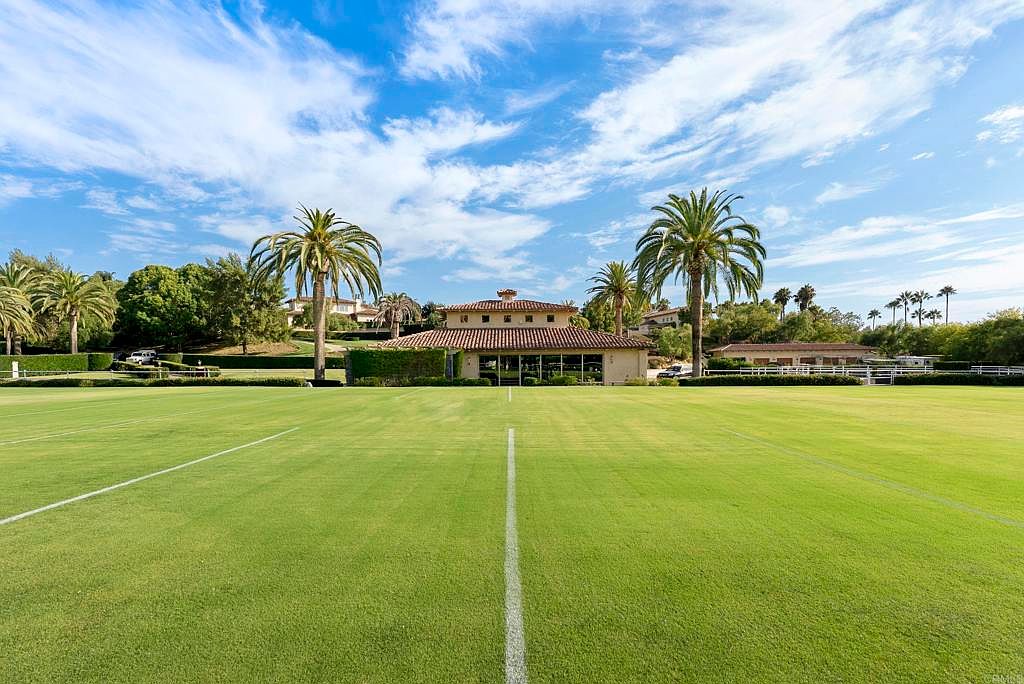 This image showcases a meticulously maintained lawn, possibly a polo field or large garden area, leading up to a building with a red tile roof and palm trees. The expansive green space is neatly striped, suggesting professional landscaping. The scene is set under a bright, partly cloudy sky, creating a serene and luxurious atmosphere.