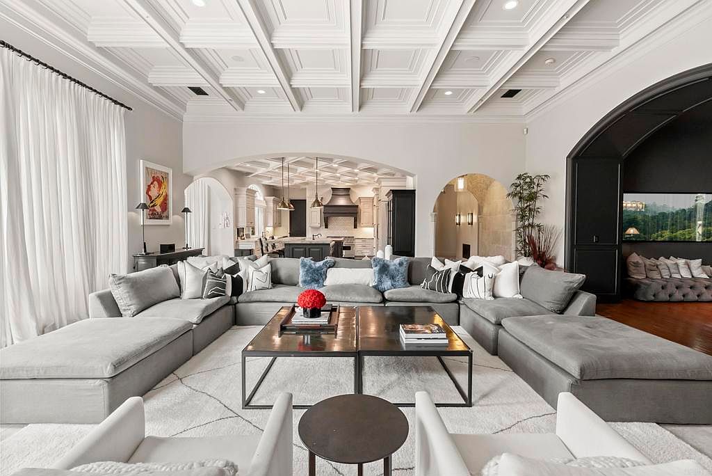 This is a spacious living room featuring a large, sectional sofa arranged around a central coffee table. The room is decorated in neutral tones with white walls, a coffered ceiling, and large windows with sheer curtains. The open floor plan leads to the kitchen and other areas of the house, creating a seamless flow.