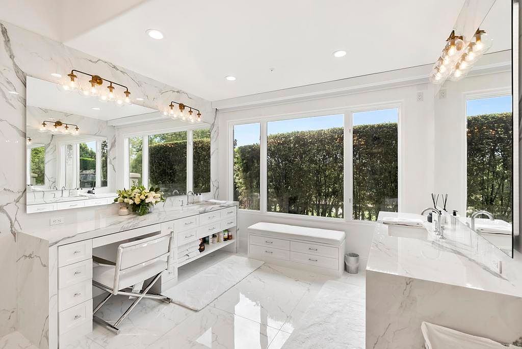 This is a luxurious primary bathroom featuring marble countertops and flooring, creating a bright and elegant space. The room includes a vanity area with a chair, ample storage, and large windows that provide natural light and a view of the greenery outside. The overall impression is one of sophistication and tranquility.