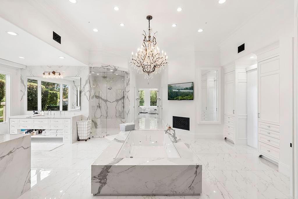 This is a luxurious primary bathroom featuring marble flooring and walls, a large soaking tub with a marble surround, and a chandelier. The bathroom also includes a glass-enclosed shower, a vanity with ample storage, and a television mounted above a fireplace. The overall impression is one of opulence and relaxation.