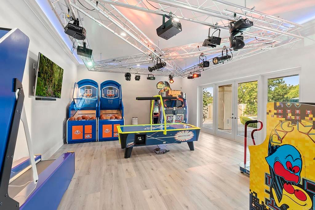 This is an interior shot of a home gym or entertainment room featuring several arcade games, including basketball hoops, an air hockey table, and a Pac-Man machine. The room is well-lit with a complex lighting system installed on the ceiling, and the light wood flooring adds a modern touch. French doors lead to an outdoor area, enhancing the room's appeal as a recreational space.