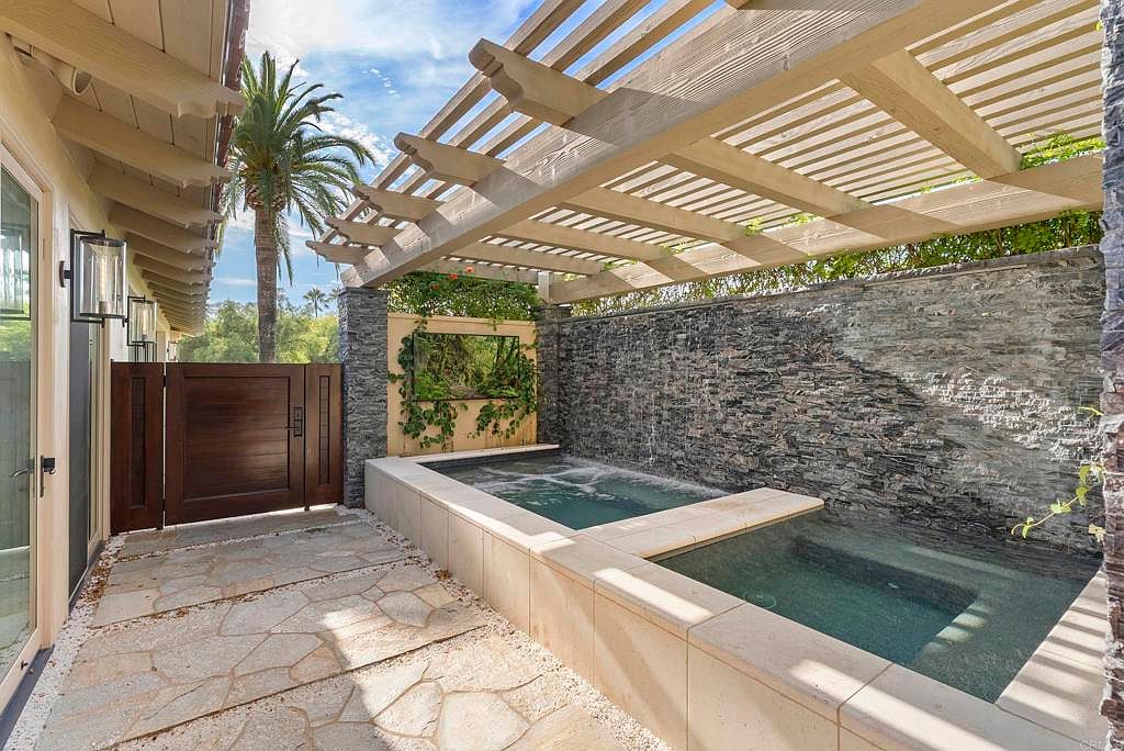 This image showcases a luxurious spa area with a stacked stone wall and a modern pergola overhead. The spa features a unique design with multiple levels and a waterfall feature, creating a tranquil and inviting atmosphere. The surrounding patio is paved with natural stone, adding to the upscale aesthetic.