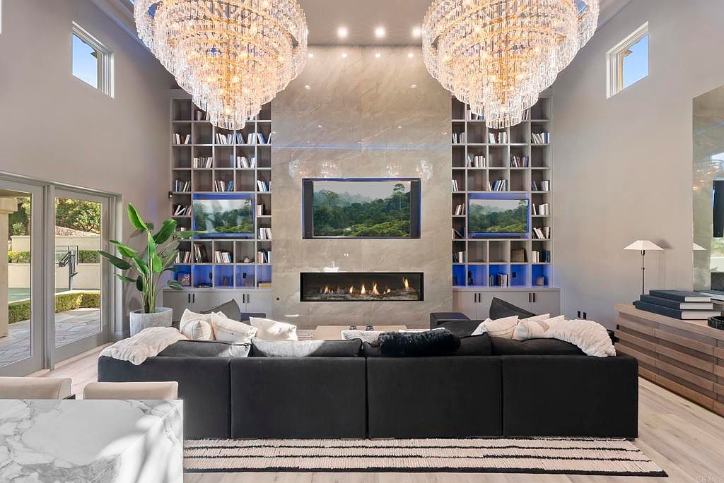 This is an interior shot of a luxurious living room featuring a large, dark sectional sofa with throw pillows and blankets, facing a modern fireplace and a wall-mounted television. Flanking the television are built-in bookshelves with accent lighting, and two large crystal chandeliers hang from the high ceiling. The room exudes a sense of opulence and comfort.