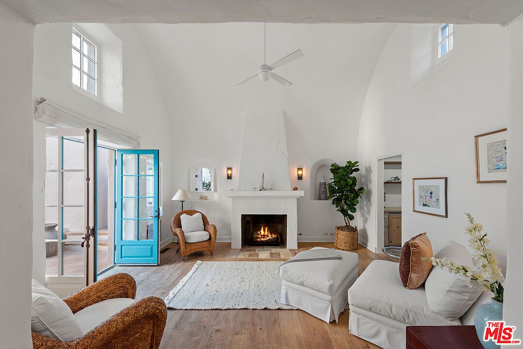 This is an interior shot of a living room featuring a white vaulted ceiling and a fireplace. The room is furnished with wicker chairs, a white ottoman, and a light-colored rug. A bright blue door leads to an outdoor space, and the overall aesthetic is bright and airy.