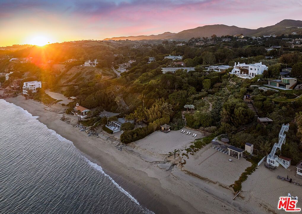 This aerial view showcases a stunning coastal property at sunset. The image captures multiple luxury homes nestled along a sandy beach, with lush greenery covering the hillside. The golden light of the setting sun adds warmth and highlights the architectural details of the residences, creating an inviting and exclusive atmosphere.