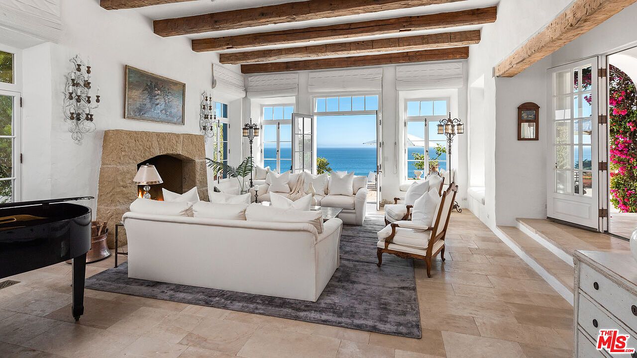 This is a bright and airy living room with a stunning ocean view. The room features exposed wooden beams, white sofas and chairs, and a stone fireplace. Large windows and doors open to the outside, creating a seamless indoor-outdoor living experience. A black piano sits to the side.