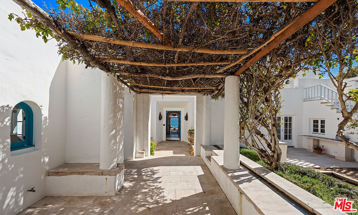 This image showcases a charming entryway featuring a pergola-covered walkway with white columns and stone flooring. The design incorporates a blend of natural elements and architectural details, creating an inviting and elegant entrance. In the background, a glimpse of the ocean adds to the property's allure.