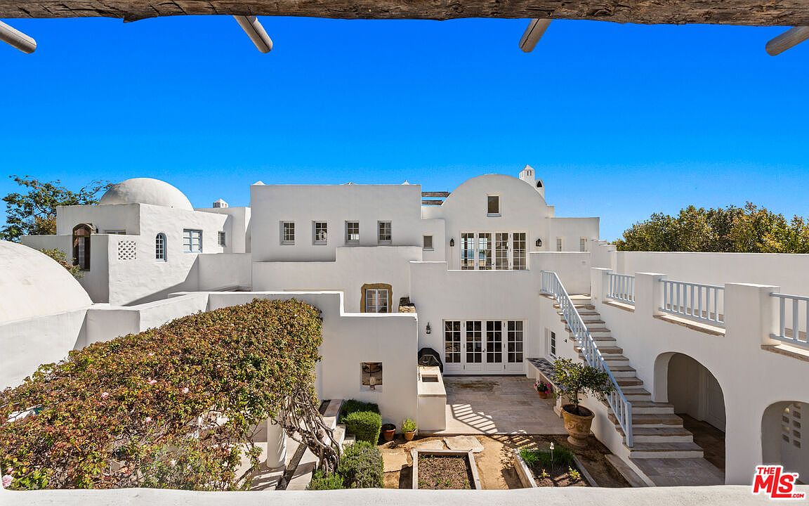 This image showcases the rear exterior of a stunning white Mediterranean-style home under a clear blue sky. The architecture features a mix of flat and domed roofs, multiple windows, and a staircase leading to an upper level. A lush garden area with mature plants and trees adds to the property's charm and privacy.
