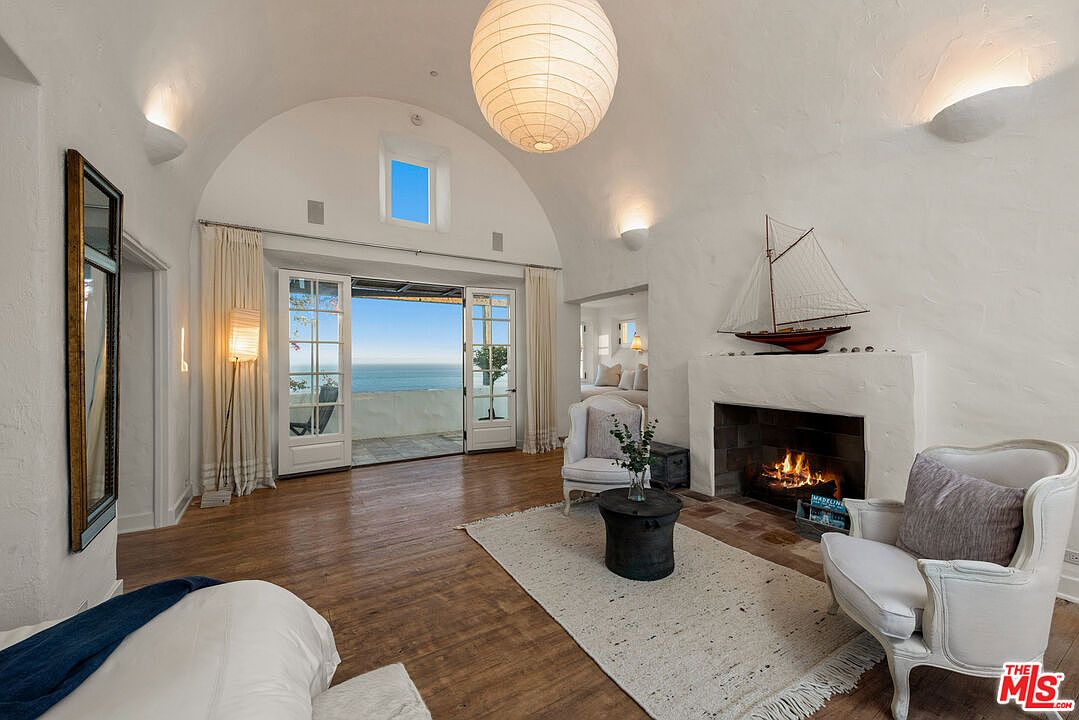 This is an interior shot of a living room featuring a barrel vaulted ceiling, a fireplace with a sailboat on the mantle, and French doors leading to a balcony with an ocean view. The room is decorated in a classic style with white armchairs, a round coffee table, and a light-colored rug. The perspective is from the back of the room, looking towards the balcony.