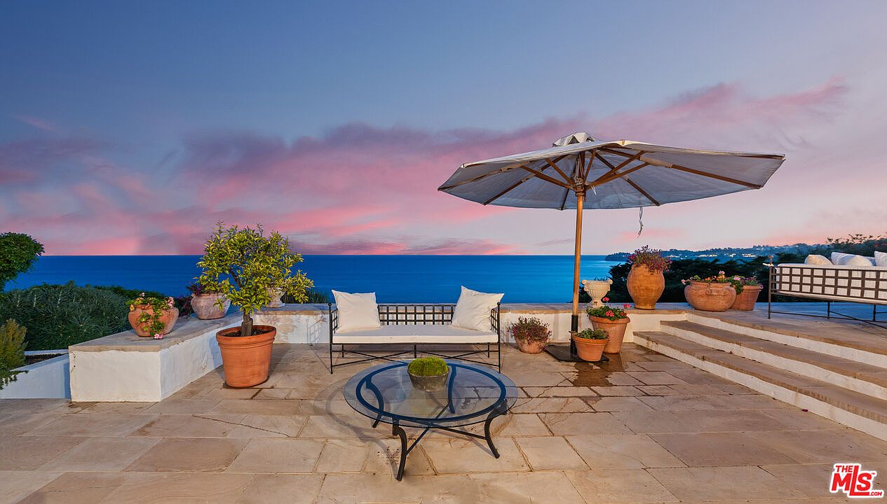 This image showcases a luxurious outdoor patio or balcony area with stunning ocean views at dusk. The space is furnished with comfortable seating, including a cushioned bench and a glass-topped table, and is adorned with potted plants and an umbrella for shade. The stone flooring and steps add to the elegant and inviting atmosphere, perfect for relaxation and enjoying the scenic surroundings.