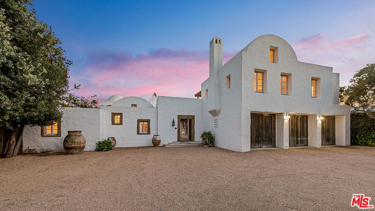 This is a front exterior view of a unique, white-washed Mediterranean-style home. The property features a gravel driveway, a prominent arched garage with wooden doors, and a charming entryway. The sky is painted with hues of pink and blue, creating a serene and inviting atmosphere.