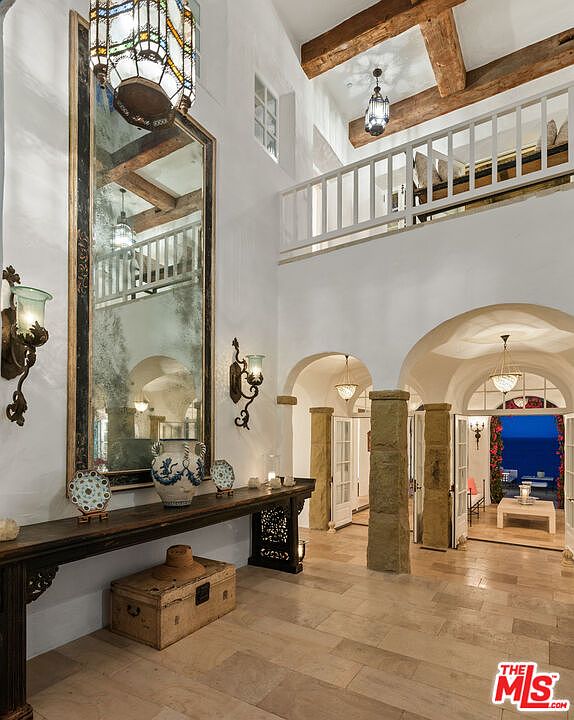 This interior shot showcases a grand hallway with high ceilings, exposed wooden beams, and a second-story balcony. The hallway features arched doorways leading to other rooms, stone pillars, and a long console table adorned with decorative items beneath a large mirror. The flooring is light-colored tile, and the overall aesthetic is luxurious and Mediterranean-inspired.