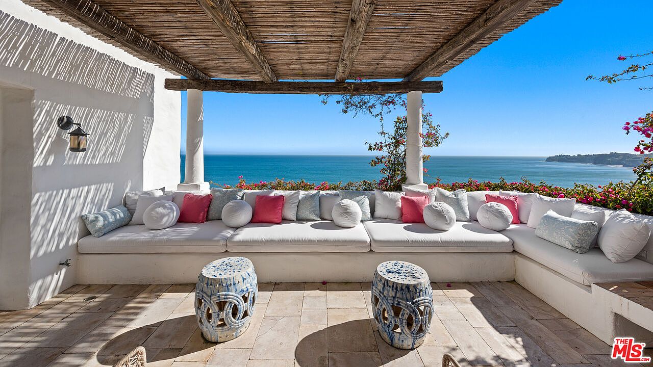 This image showcases a luxurious outdoor patio or balcony with stunning ocean views. A large, built-in white cushioned seating area is adorned with an array of decorative pillows in various colors and patterns. Two decorative ceramic stools sit in front of the seating, and a rustic wooden pergola provides shade, creating an inviting and relaxing outdoor living space.
