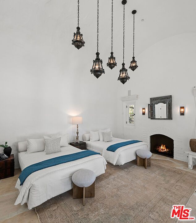 This interior shot showcases a guest bedroom featuring two twin beds with white linens and blue accents. The room is illuminated by unique hanging light fixtures and a table lamp, creating a warm ambiance. A fireplace adds a cozy touch, while the overall style is clean and inviting.