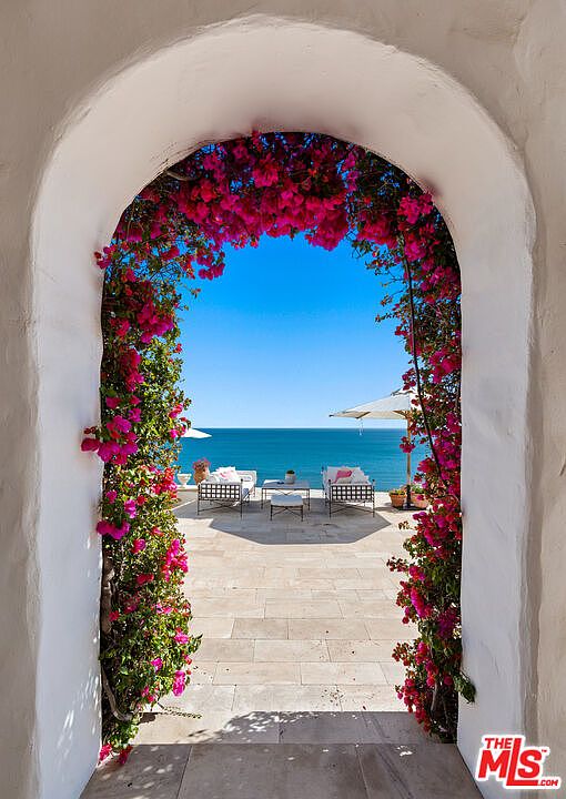 This image showcases a beautiful patio or balcony with an ocean view, framed by a white arched doorway adorned with vibrant pink bougainvillea. The patio features tiled flooring and outdoor furniture, including lounge chairs and a small table, inviting relaxation and enjoyment of the coastal scenery. The clear blue sky and ocean create a serene and luxurious atmosphere.