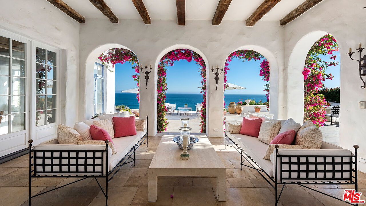 This image showcases a stunning outdoor patio or balcony area with a breathtaking ocean view framed by arched openings adorned with vibrant pink bougainvillea. The space is furnished with two elegant daybeds featuring black metal frames and light-colored cushions, flanking a simple stone coffee table. The overall aesthetic is luxurious and inviting, perfect for relaxation and enjoying the coastal scenery.