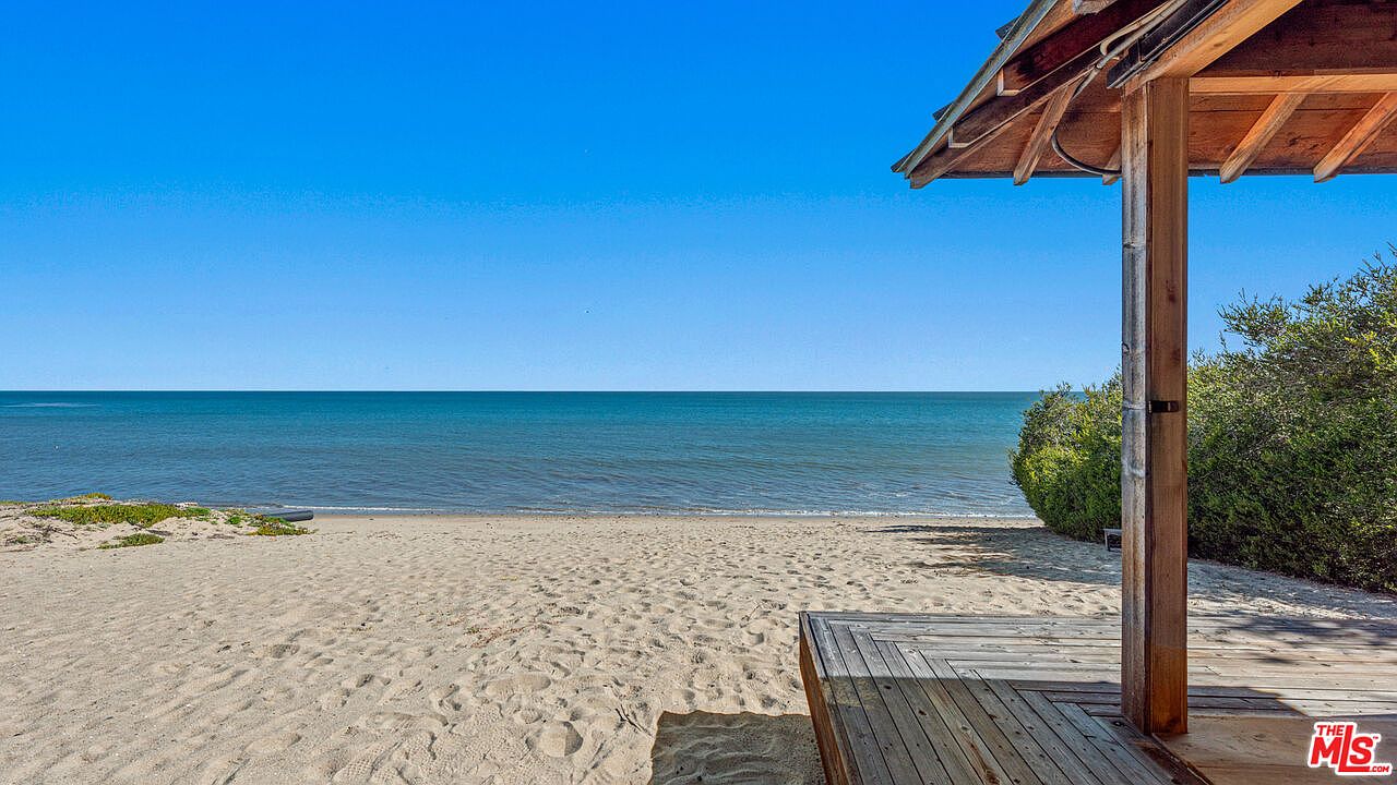 This image showcases a serene beachside patio or deck, offering direct access to a sandy beach and the ocean. The wooden structure provides a shaded area, while the deck extends towards the beach, creating a seamless transition between the built environment and the natural landscape. The clear blue sky and calm water enhance the tranquil atmosphere, making it an ideal spot for relaxation and enjoying the coastal scenery.