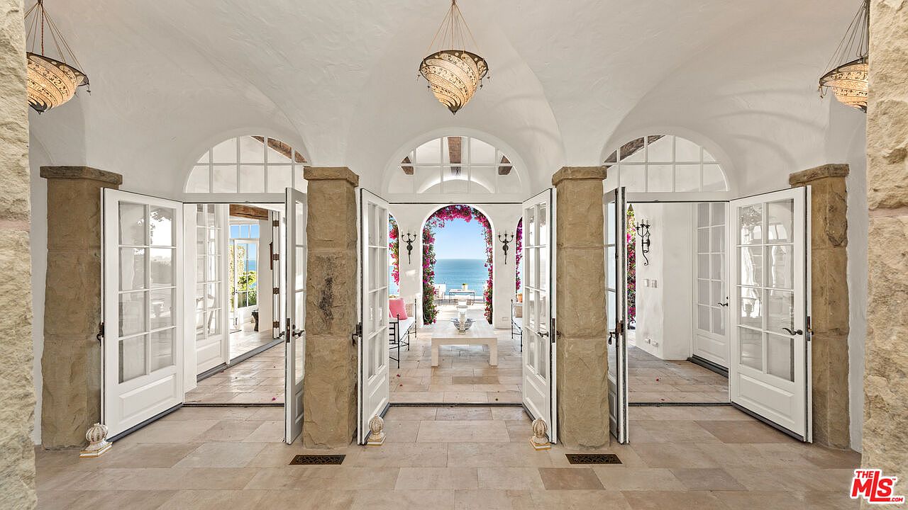This interior shot showcases a grand hallway with a series of arched doorways leading to an outdoor patio with ocean views. The hallway features stone pillars, white-framed glass doors, and a light-colored tiled floor, creating a bright and airy atmosphere. The design emphasizes a seamless transition between indoor and outdoor living spaces.