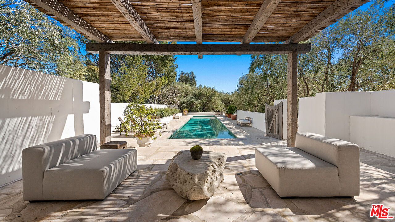 This image showcases a luxurious outdoor living space featuring a rectangular swimming pool as the focal point. The pool is surrounded by a stone patio furnished with modern sofas and a unique stone coffee table. A pergola provides shade, creating an inviting and relaxing atmosphere, perfect for entertaining or unwinding.