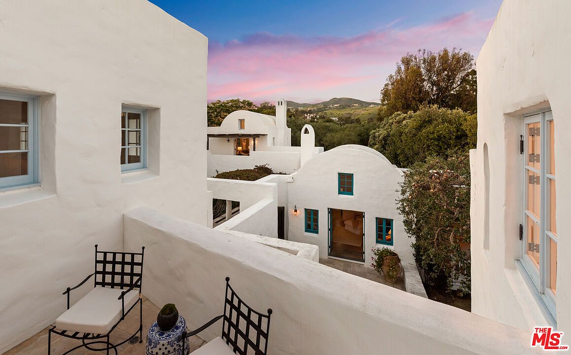 This image showcases a charming patio or balcony area with white stucco walls and blue-framed windows, evoking a Mediterranean or Santorini-inspired architectural style. Two black wrought-iron chairs with white cushions are arranged on the patio, suggesting a relaxing outdoor space. The view extends to other white buildings with rounded roofs and lush greenery in the background, creating a serene and picturesque setting.
