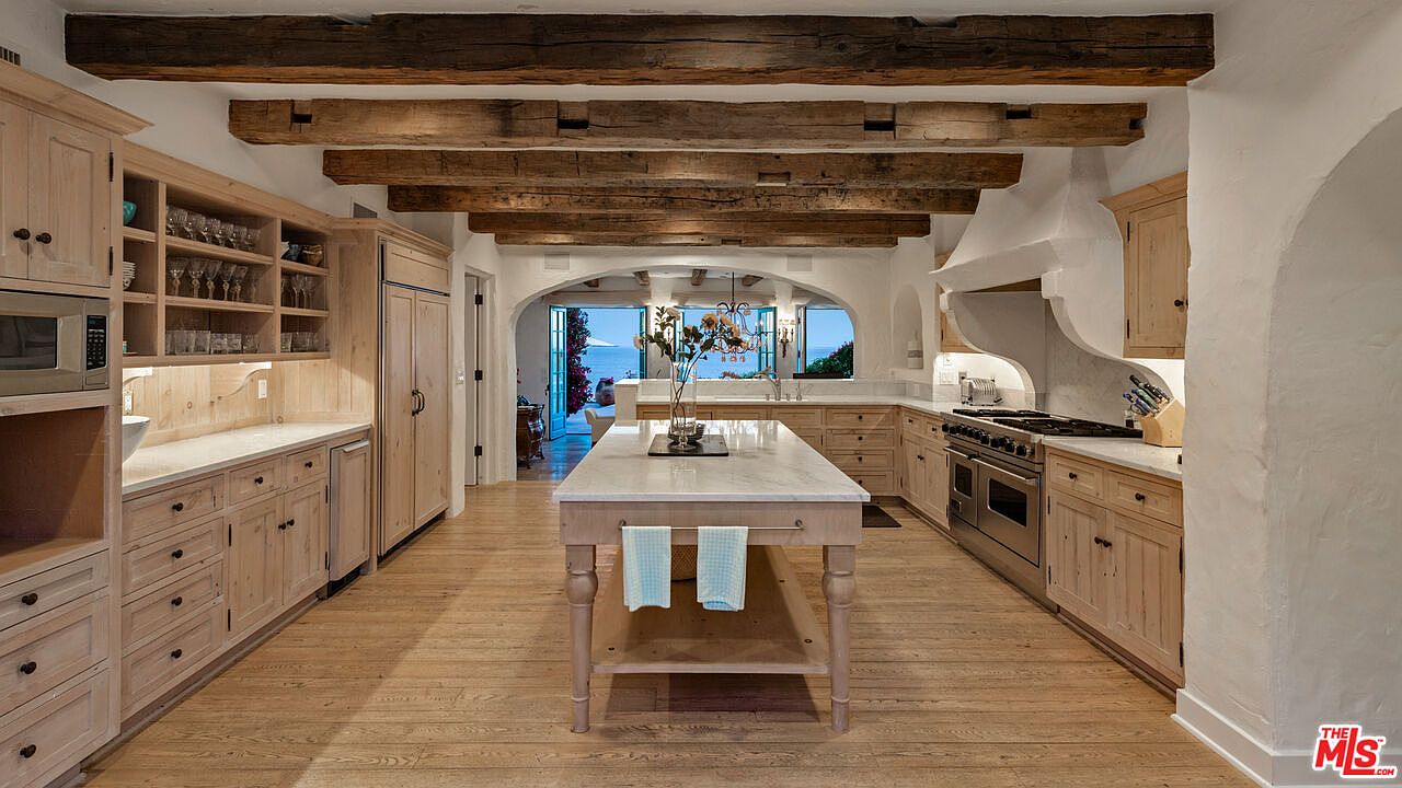 This is a spacious kitchen featuring light wood cabinetry, marble countertops, and rustic wooden beams across the ceiling. A large island with a marble top sits in the center, and an arched opening provides a view to the outside. The kitchen is well-lit and has a warm, inviting atmosphere.