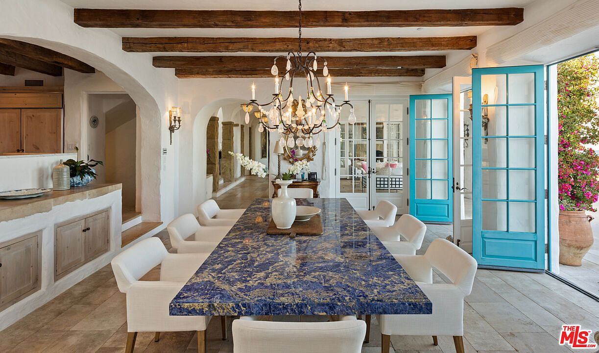 This is an interior shot of a dining room featuring a long, blue marble-like table with white chairs. A chandelier hangs above the table, and the room has wooden beams on the ceiling and white walls with arched doorways. The room opens to an outdoor area through glass doors, creating a bright and airy atmosphere.