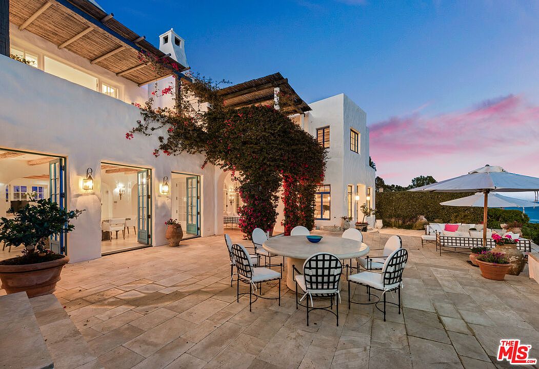 This image showcases a beautifully designed patio area of a Mediterranean-style home at dusk. The patio features a stone tile floor, a round dining table with chairs, and an outdoor seating area with umbrellas in the background. The building is adorned with climbing plants, and the open doors reveal glimpses of the interior, creating an inviting and luxurious outdoor living space.